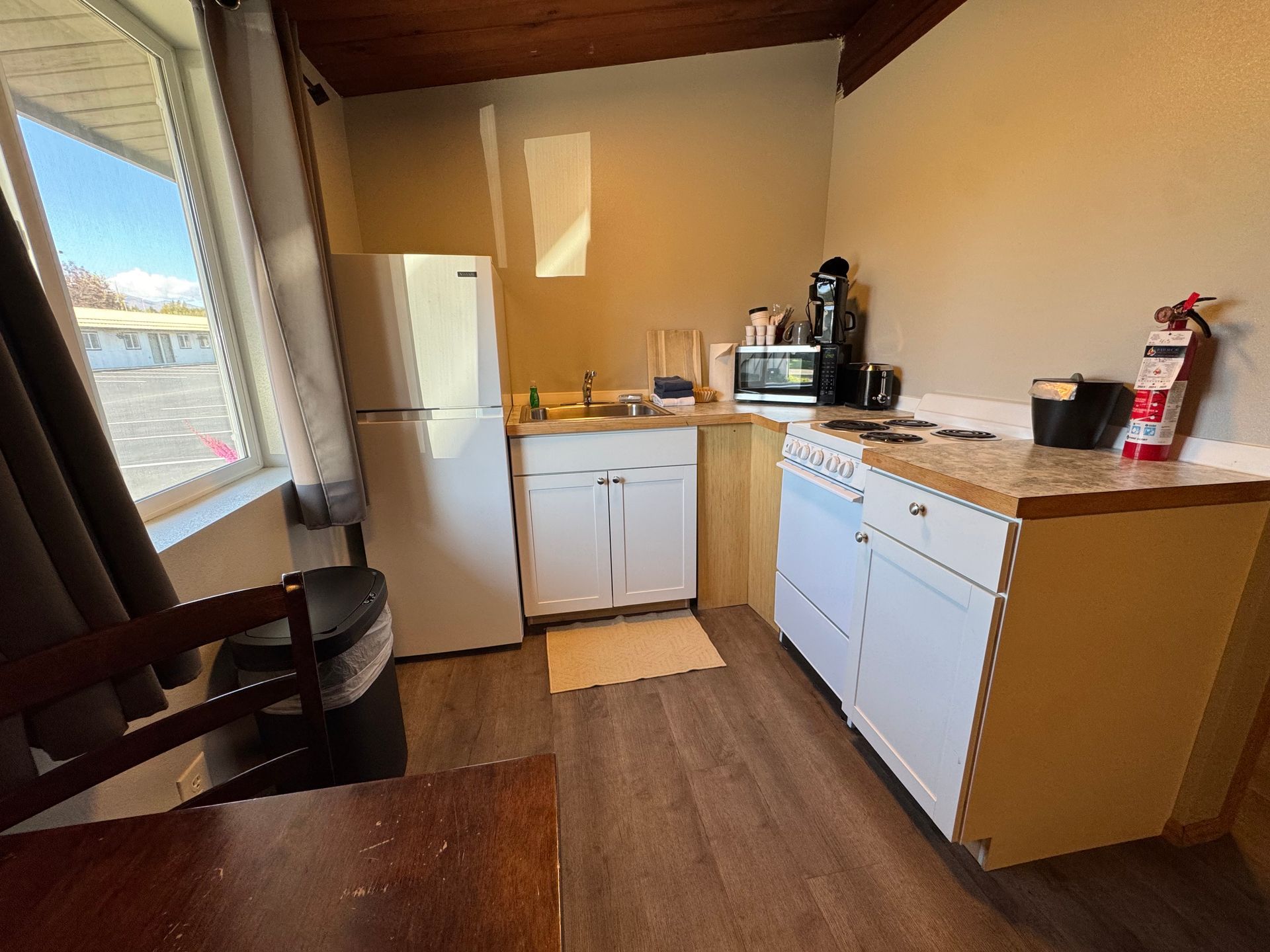 Small kitchen with white fridge, cabinets, and appliances; window on the left; fire extinguisher on the right.