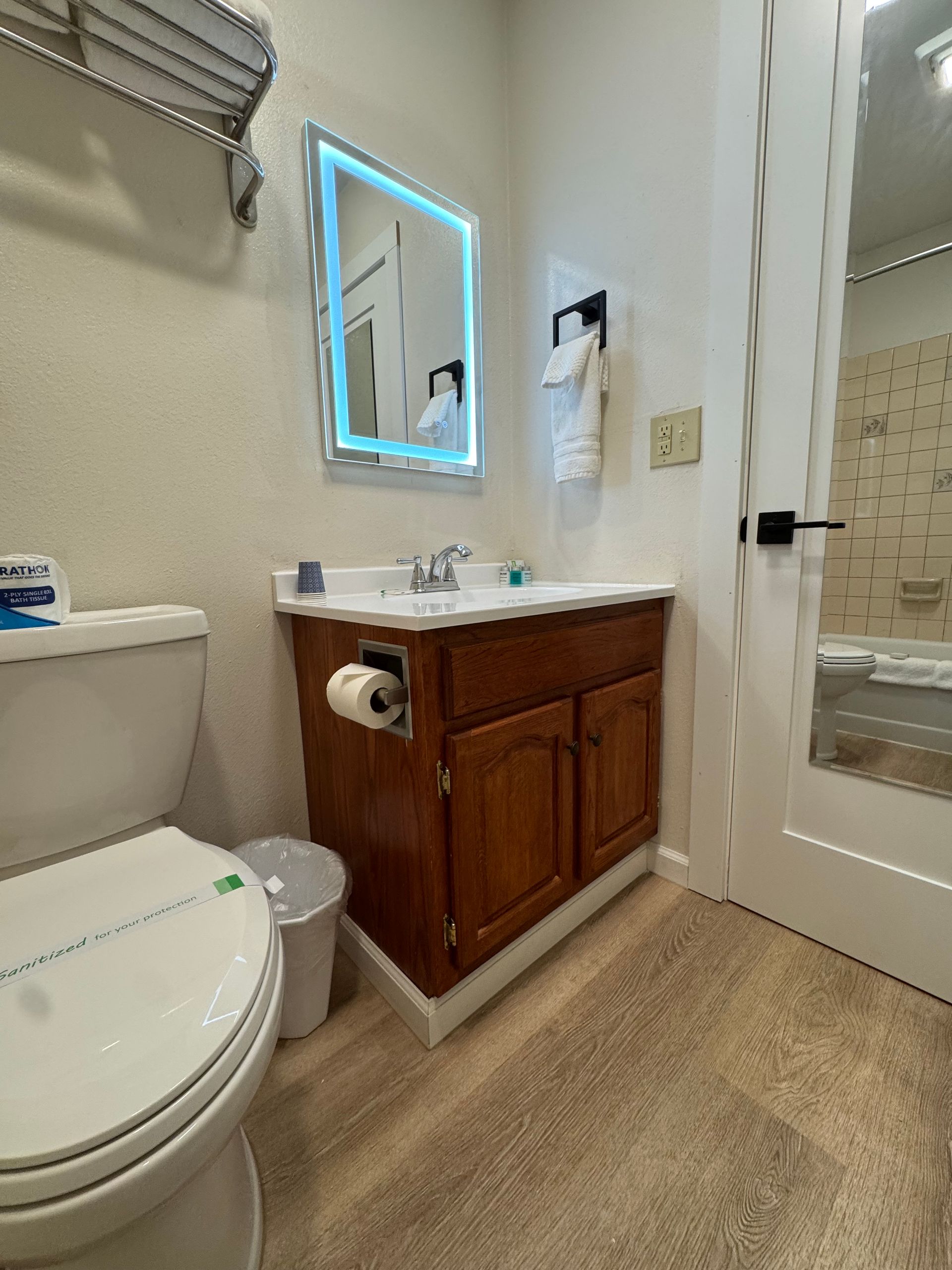 Bathroom with toilet, sink, and mirror; brown cabinets, white walls, and light brown flooring.