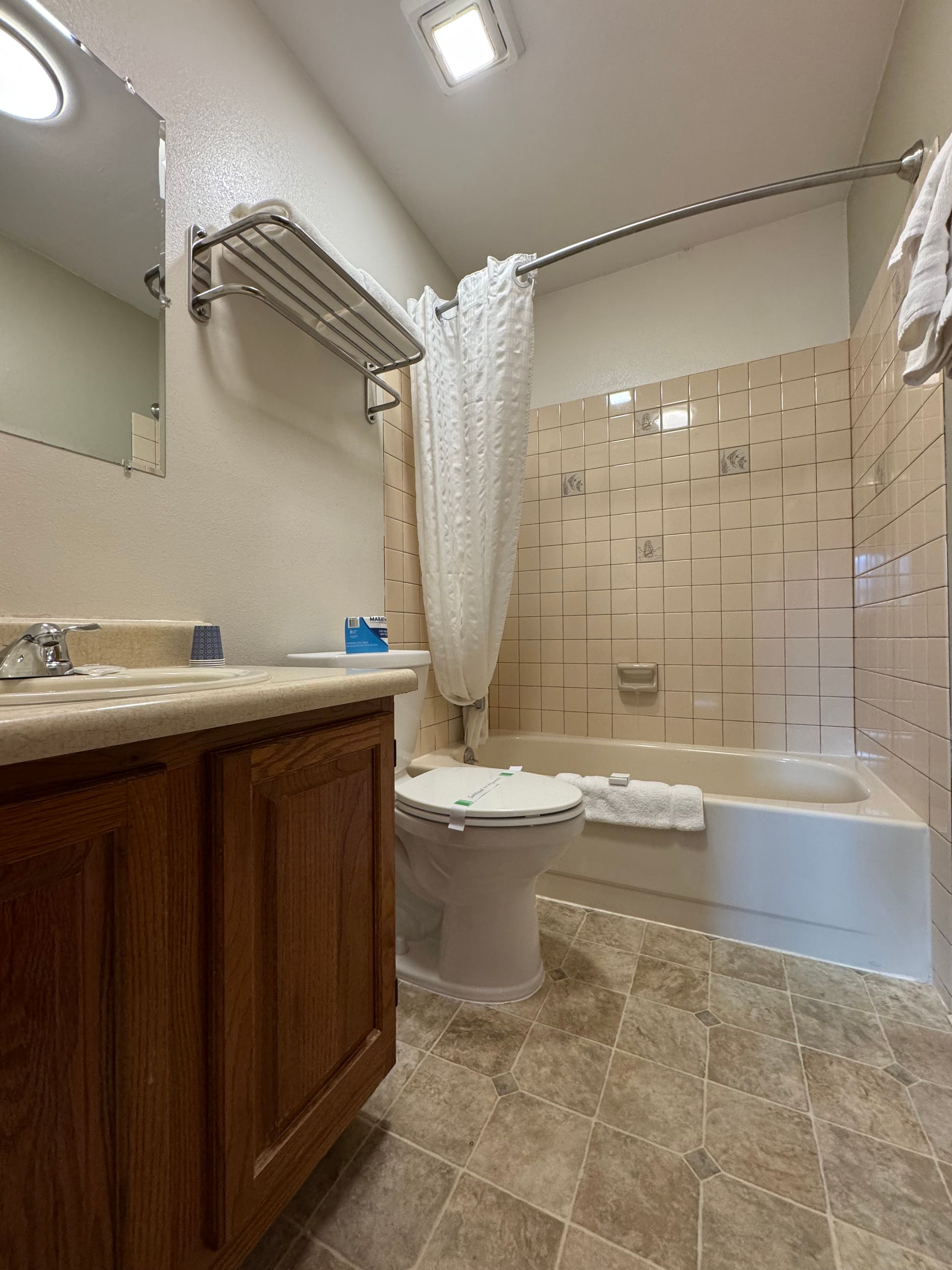 Bathroom with toilet, bathtub, and vanity. Brown cabinets and tiled floor. White shower curtain.