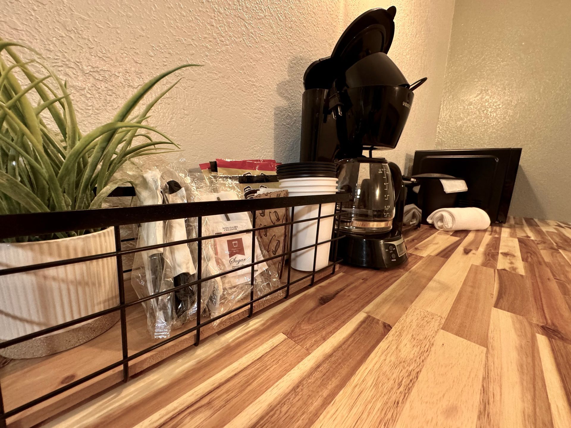 Coffee station with black coffee maker, wire rack, plant, and wooden countertop.