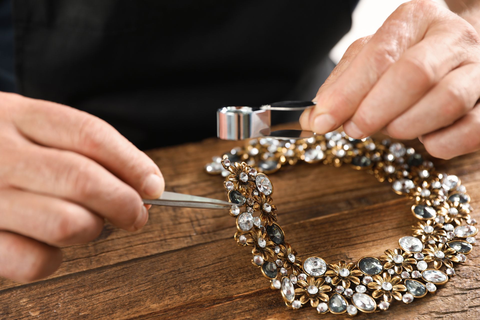 Hands using tweezers to arrange a jeweled necklace on a wooden table.