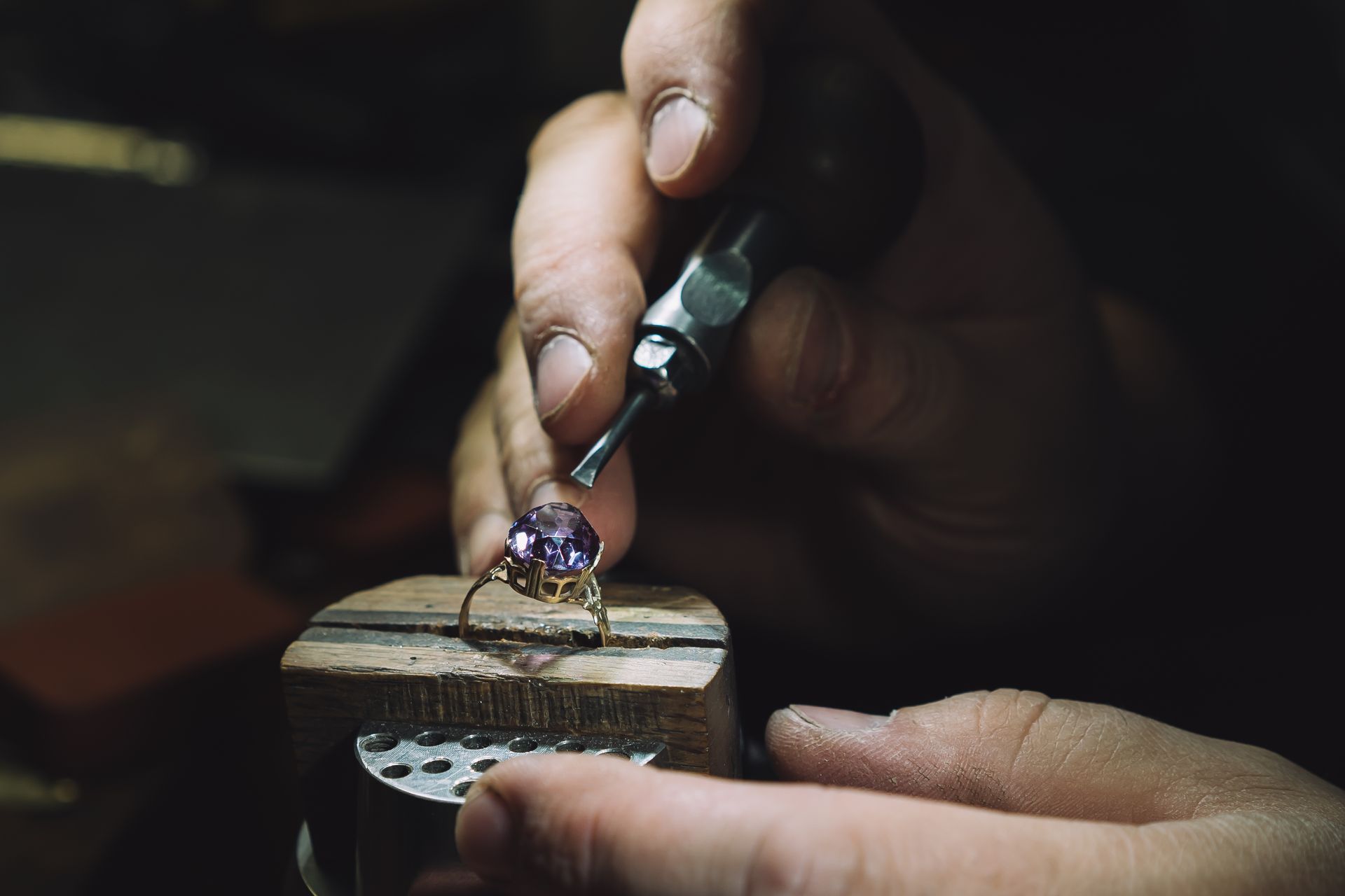 Hands using a tool to shape a small gemstone on a jeweler’s workbench