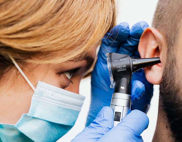 Doctor examining a patient’s ear with an otoscope, wearing a face mask and blue gloves.