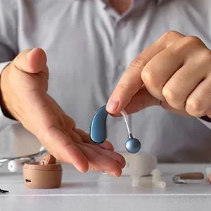 Hand demonstrating blue and silver hearing aid parts over a table with a small beige case