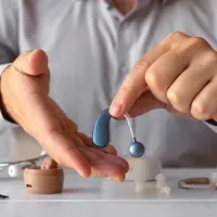 Hand demonstrating blue and silver hearing aid parts over a table with a small beige case