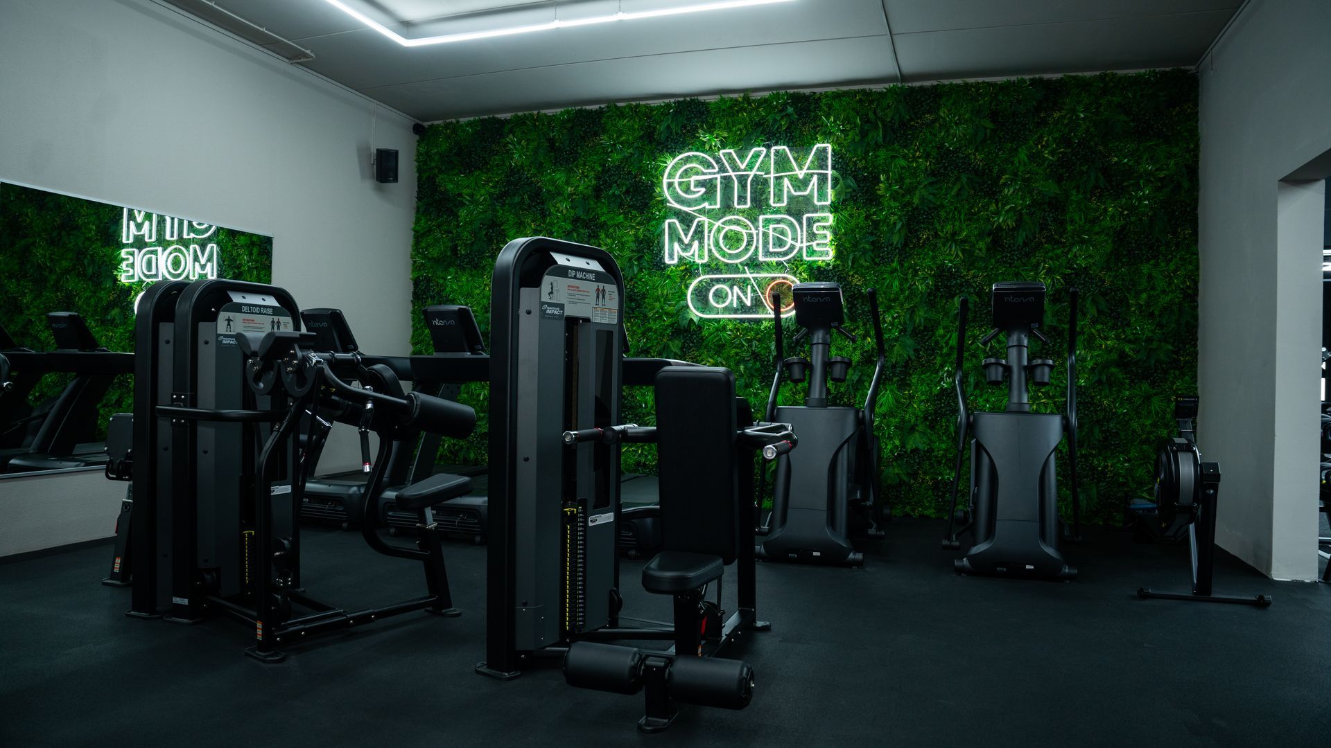Interieur van een sportschool met fitnessapparaten tegen een groene plantenwand met een neonbord met de tekst