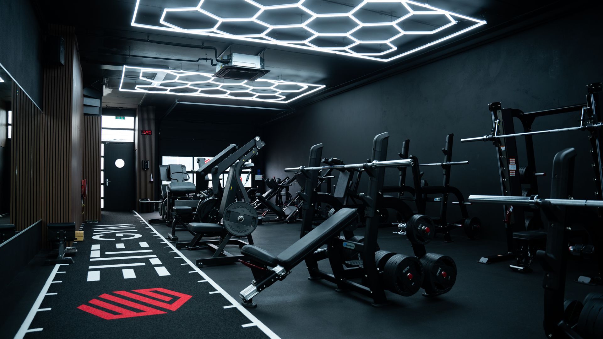 Interieur van een sportschool met fitnessapparatuur, waaronder gewichtsbanken, en zeshoekige plafondlampen.