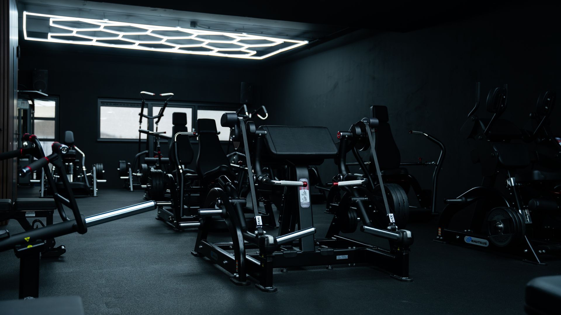 Donker, modern fitnesscentrum met gewichtsapparaten. Plafondlampen met een zeshoekig patroon verlichten de ruimte.