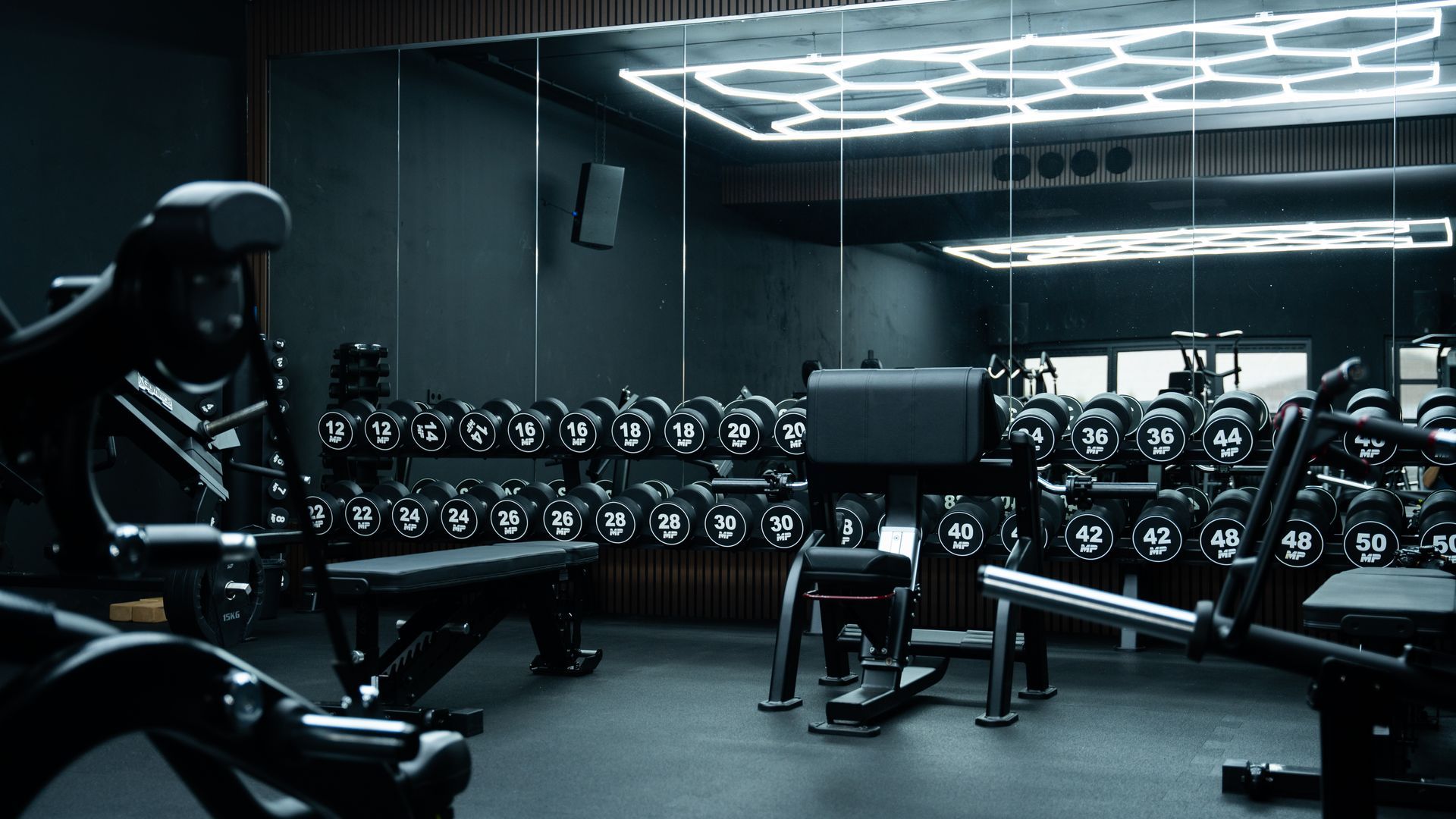 Interieur van een moderne sportschool met gewichten, banken en apparaten, overwegend donker met koele verlichting.