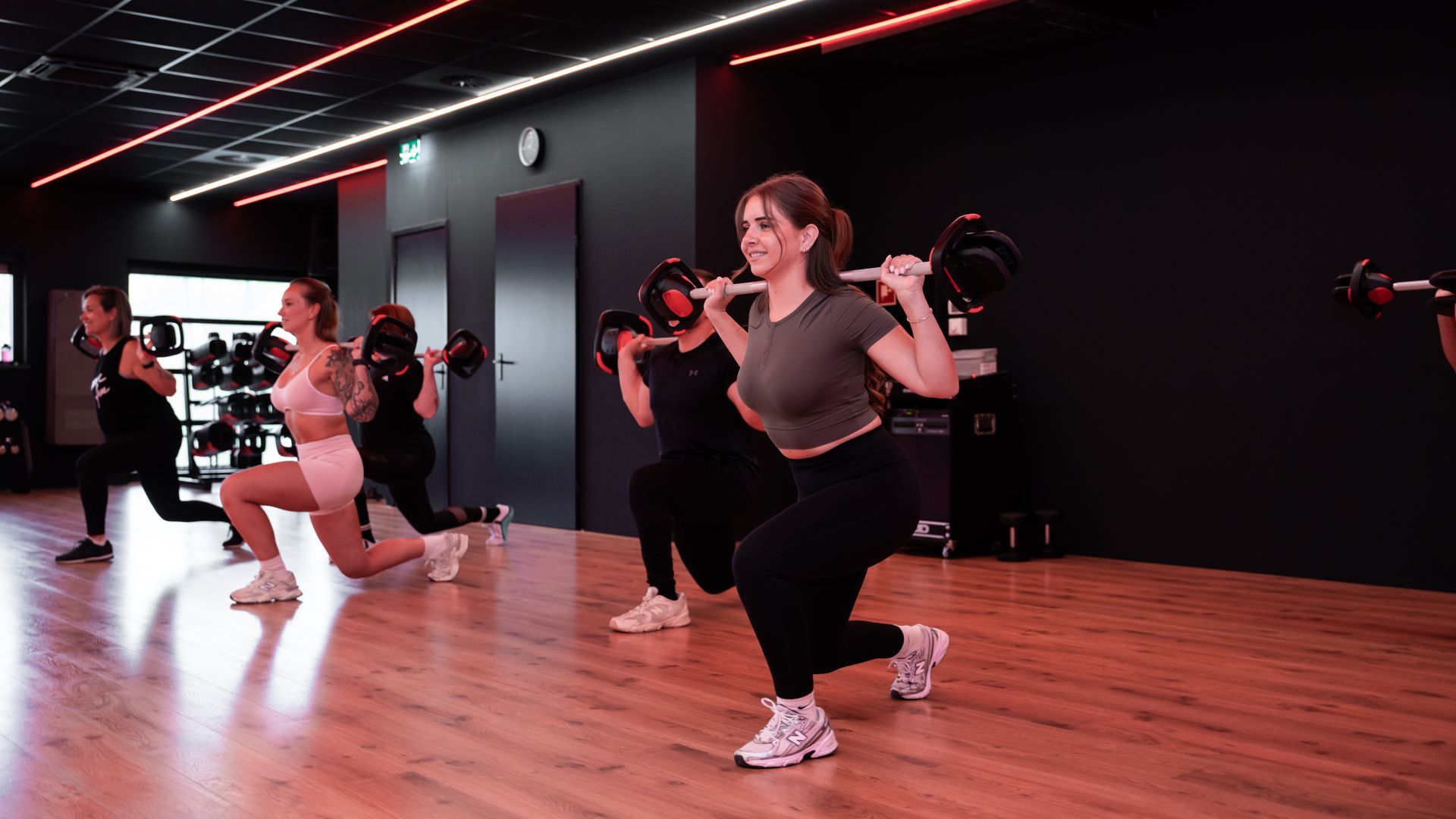 Mensen in een sportschool die lunges met gewichten uitvoeren, onder rood en wit licht.