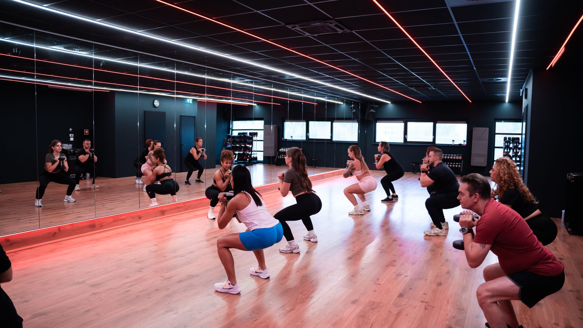 Mensen doen squats voor een spiegel tijdens een fitnessles, binnen met gekleurd licht.