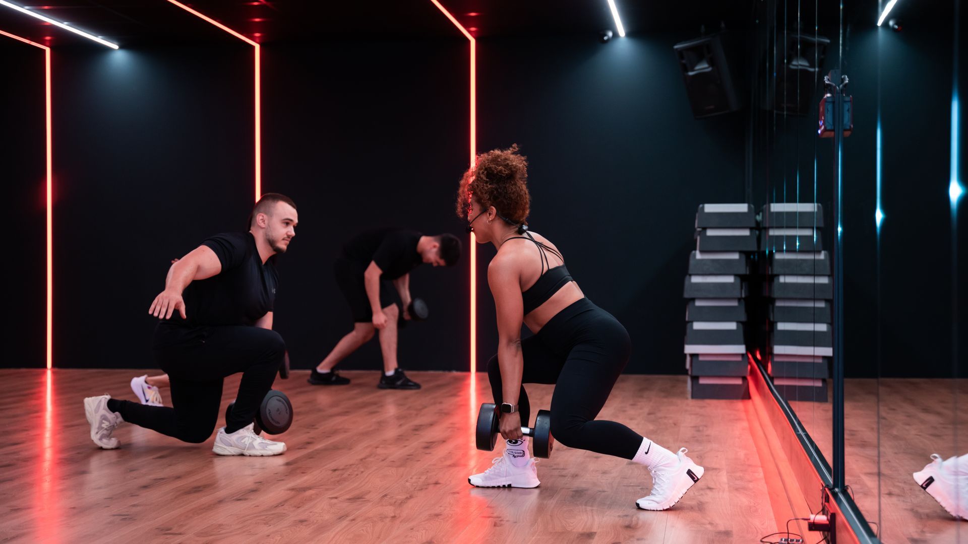 Mensen trainen in een sportschool; een trainer begeleidt iemand bij lunges met dumbbells, anderen trainen op de achtergrond.