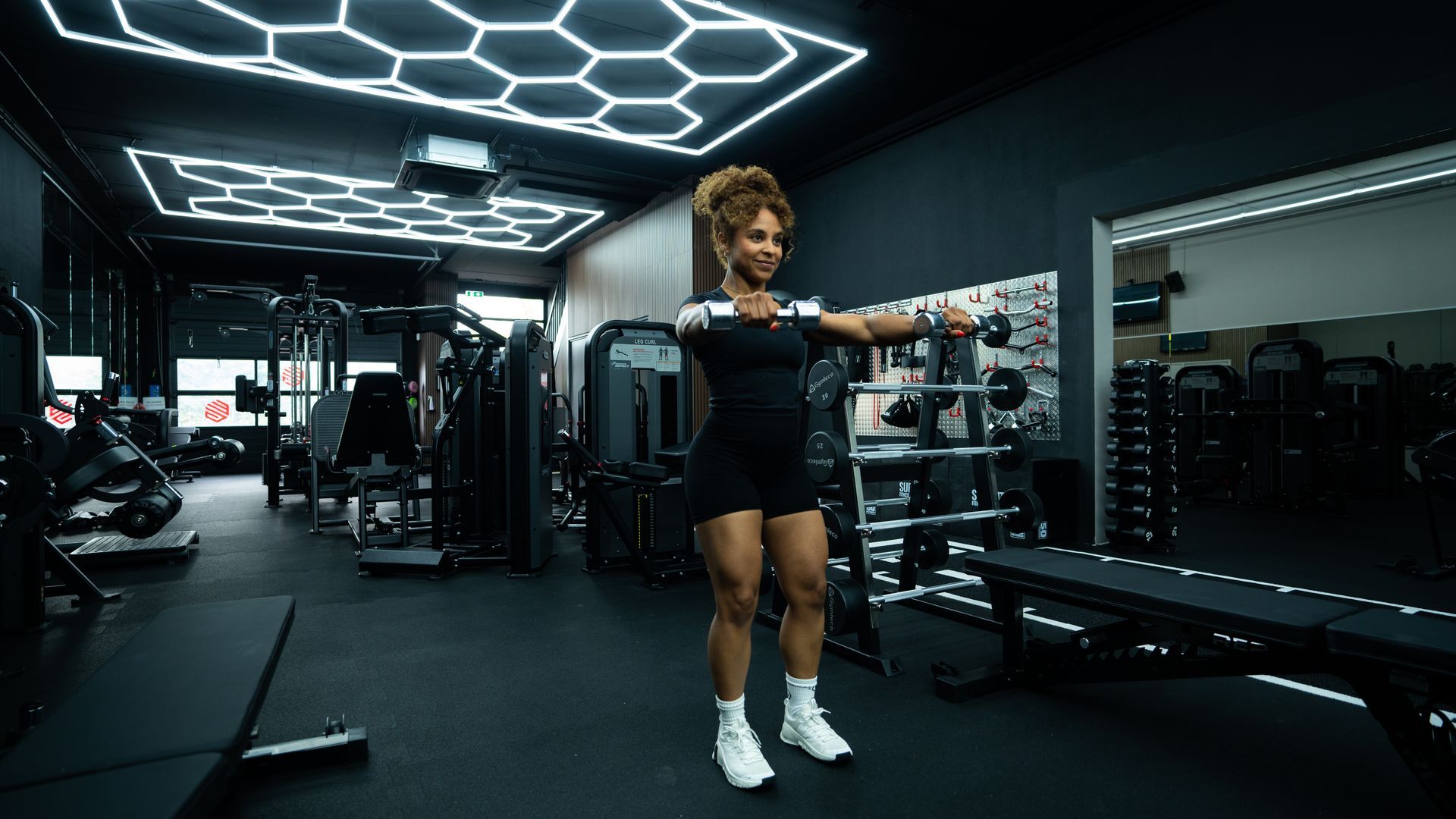 Vrouw in de sportschool met uitgestrekte armen aan het sporten. Zwarte outfit, witte schoenen. Donkere binnenruimte, fitnessapparatuur zichtbaar.