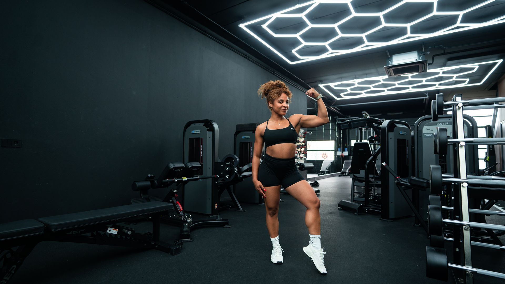 Een vrouw spant haar biceps aan in een moderne sportschool. Ze draagt zwarte sportkleding en witte sneakers, in een donkere ruimte met zeshoekige lampen.