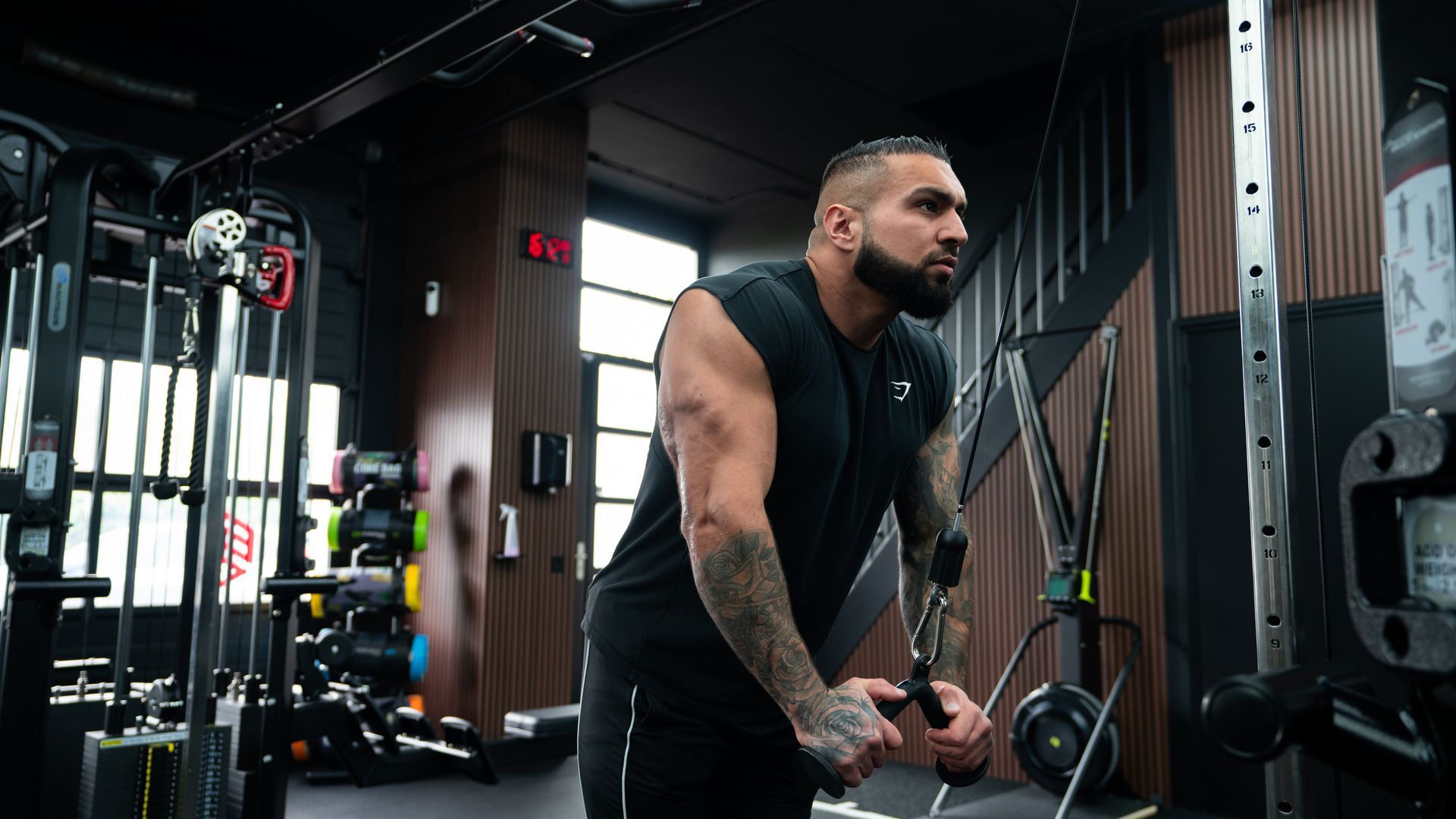 Een man is aan het trainen in de sportschool en doet triceps pushdowns op een kabelmachine. Hij draagt zwarte kleding en heeft tatoeages.