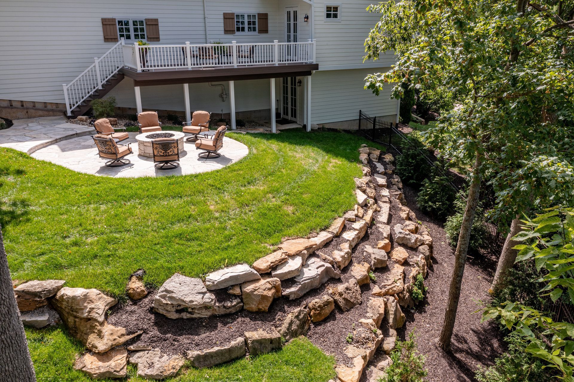 Backyard patio with fire pit, stone retaining walls, and a deck behind a white house
