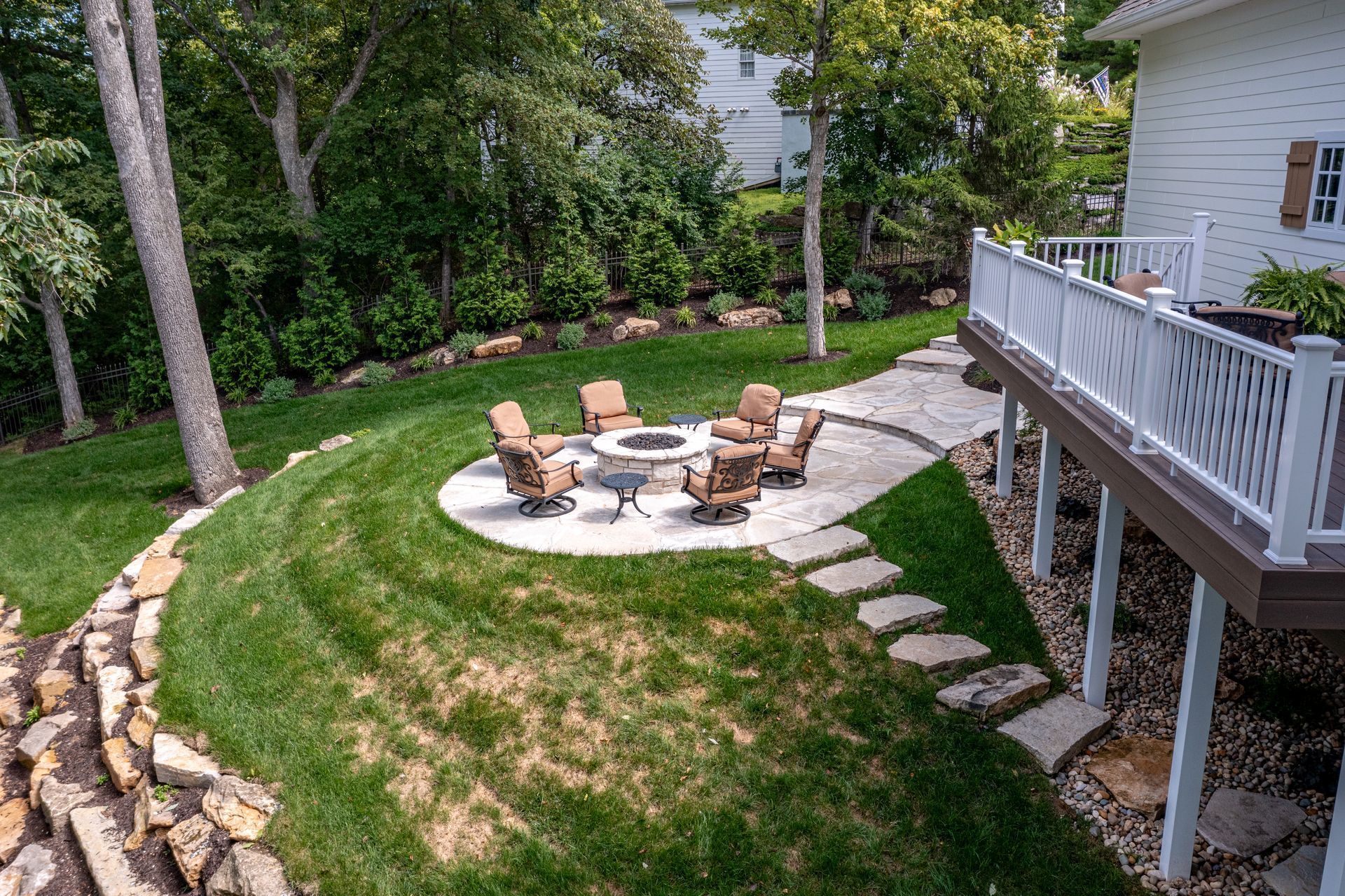 Backyard patio with fire pit, chairs, stone path, and a deck beside a grassy slope
