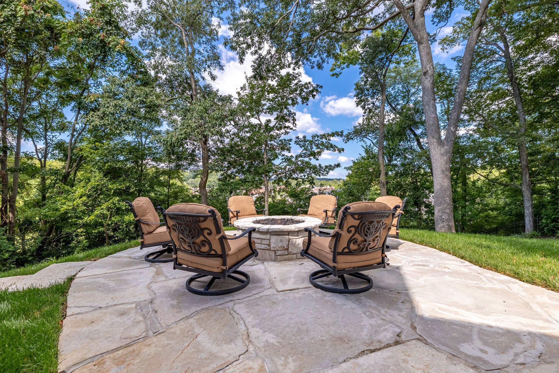 Outdoor patio with stone fire pit and six brown chairs amid trees and greenery