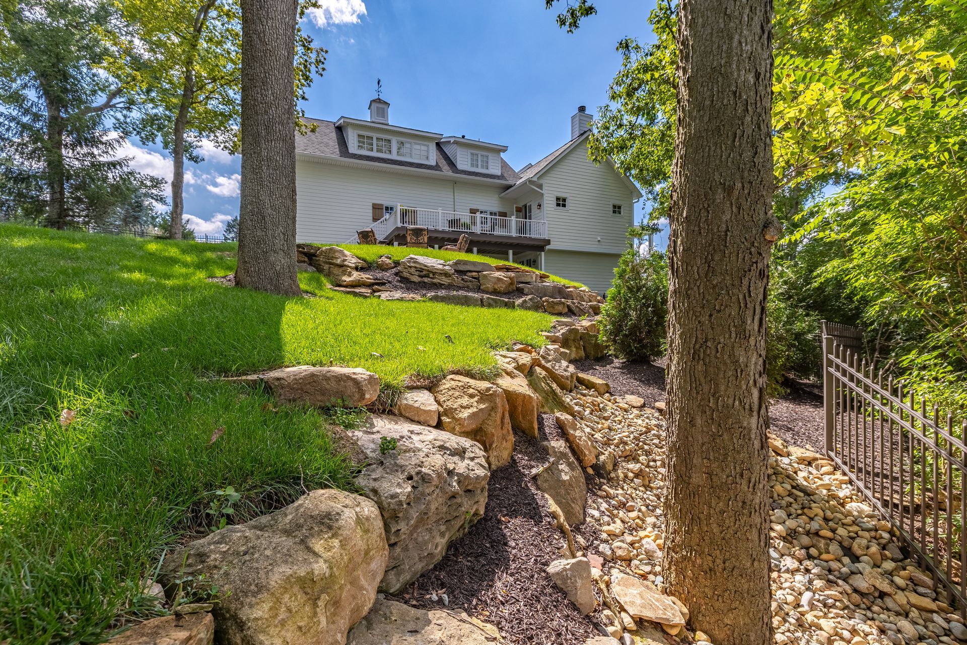 White house on a grassy hillside with stone steps and tall trees in bright sunlight