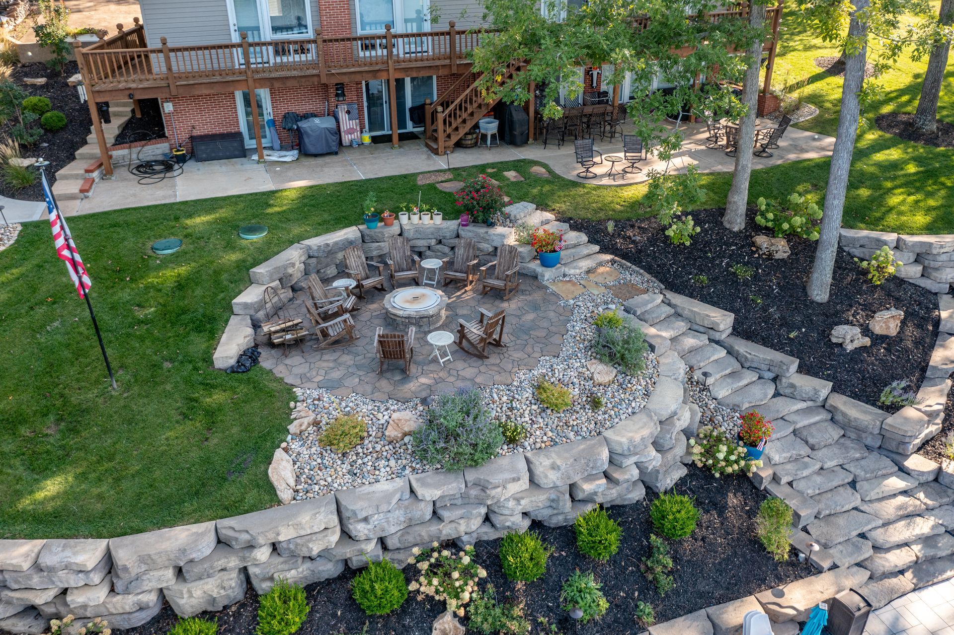 Stone fire pit surrounded by landscaping, with a wooden deck and American flag in the yard