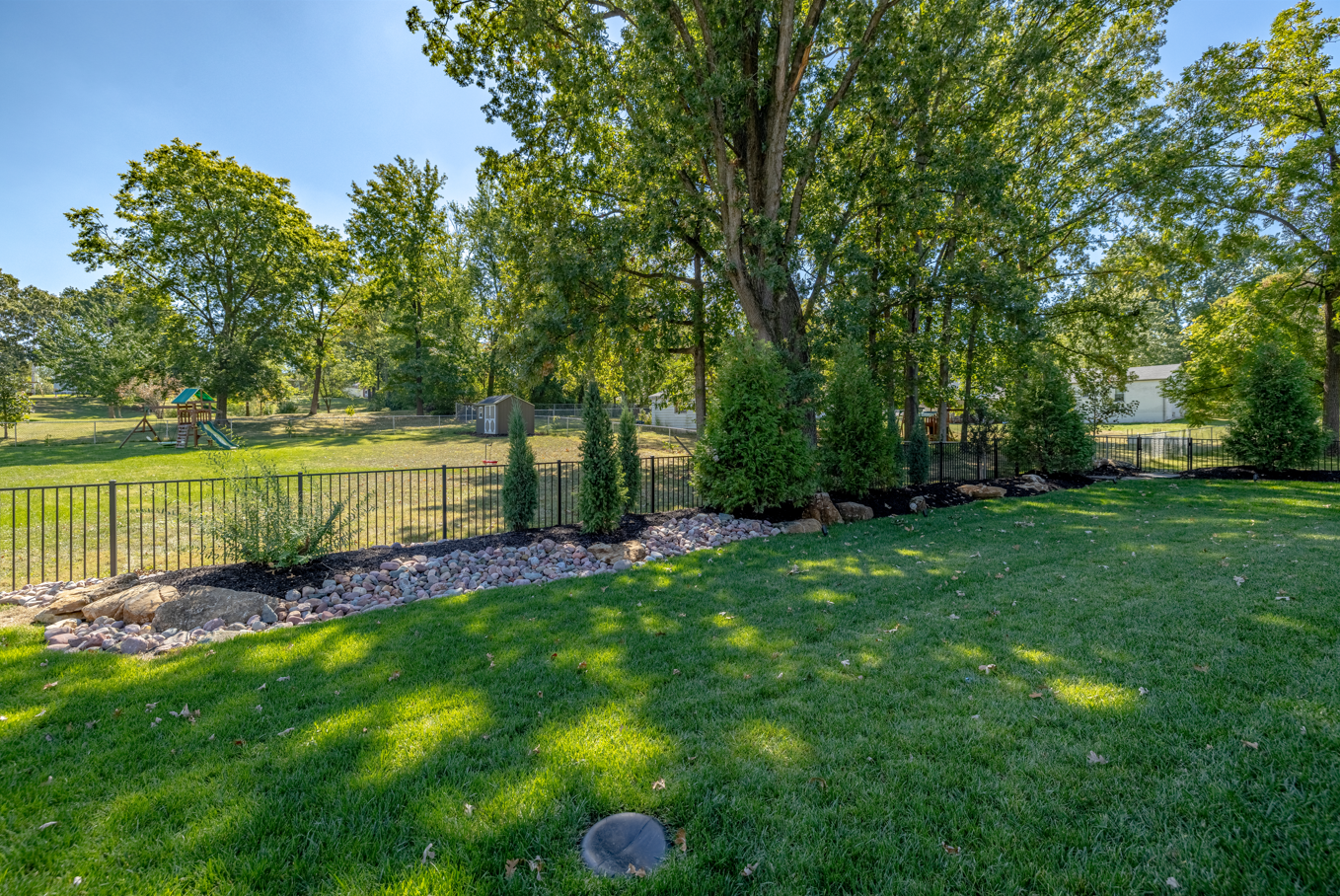 Sunny park with green lawn, tall trees, and a small fenced stream or drainage area.