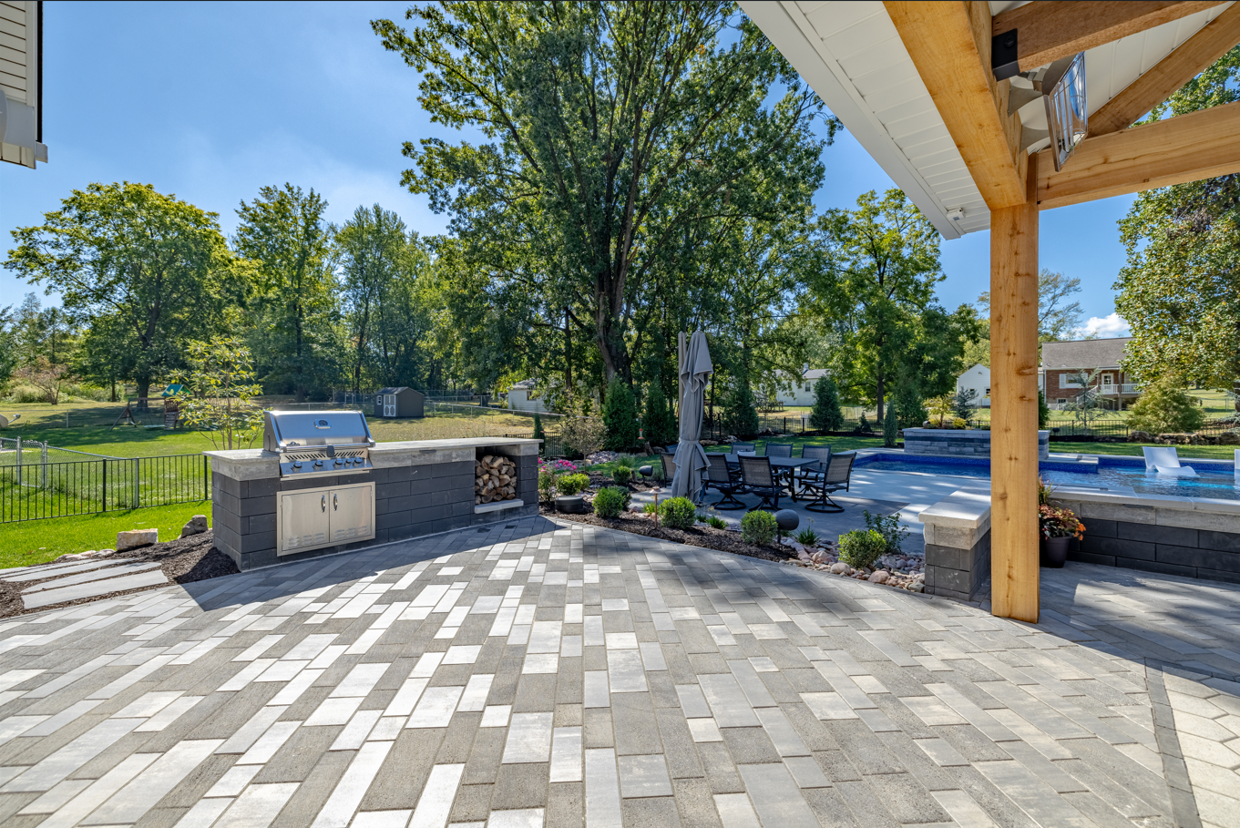 Back patio with gray pavers, built-in grill, trees, and a pool in the background