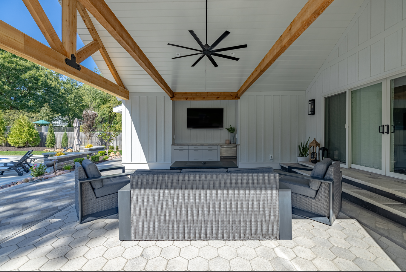 Covered patio with a TV, ceiling fan, gray seating, and a patterned bar counter overlooking a driveway.