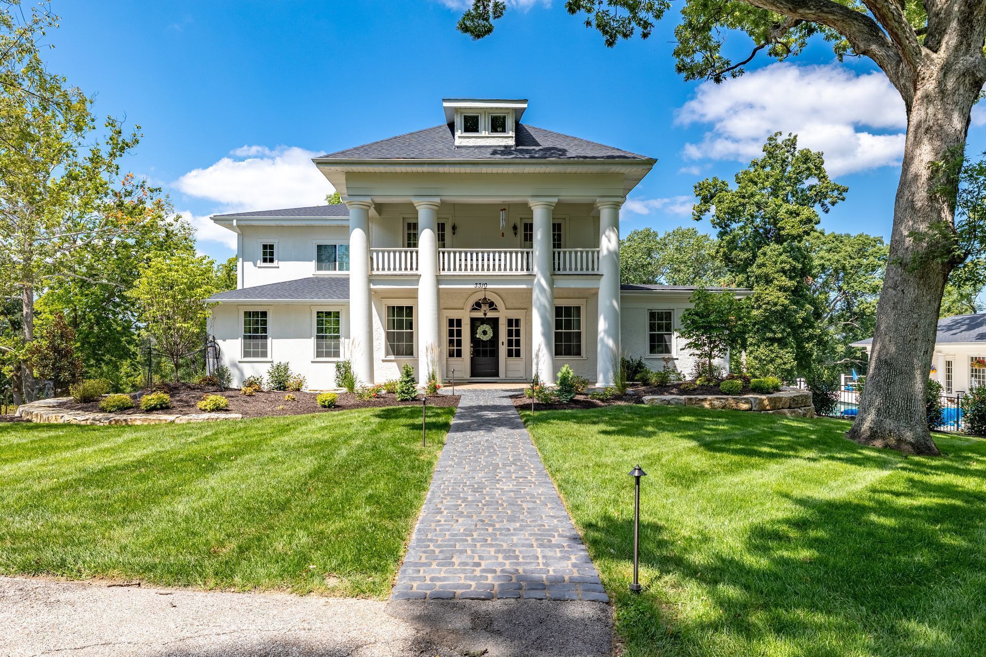 Large white colonial-style mansion with columns, front lawn, and a stone walkway under a blue sky