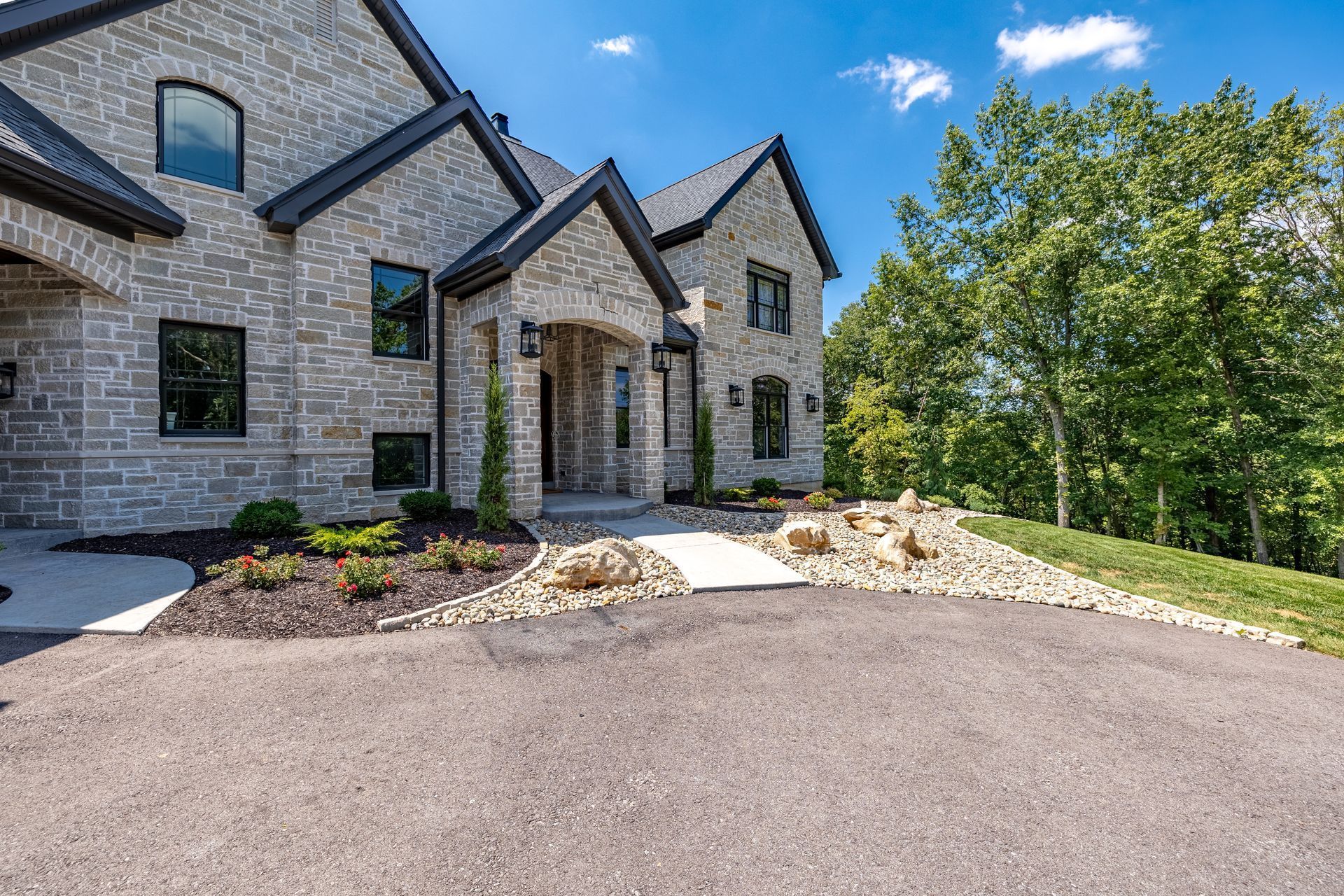 Stone house with dark trim, landscaped front entrance, and a paved driveway under a clear blue sky