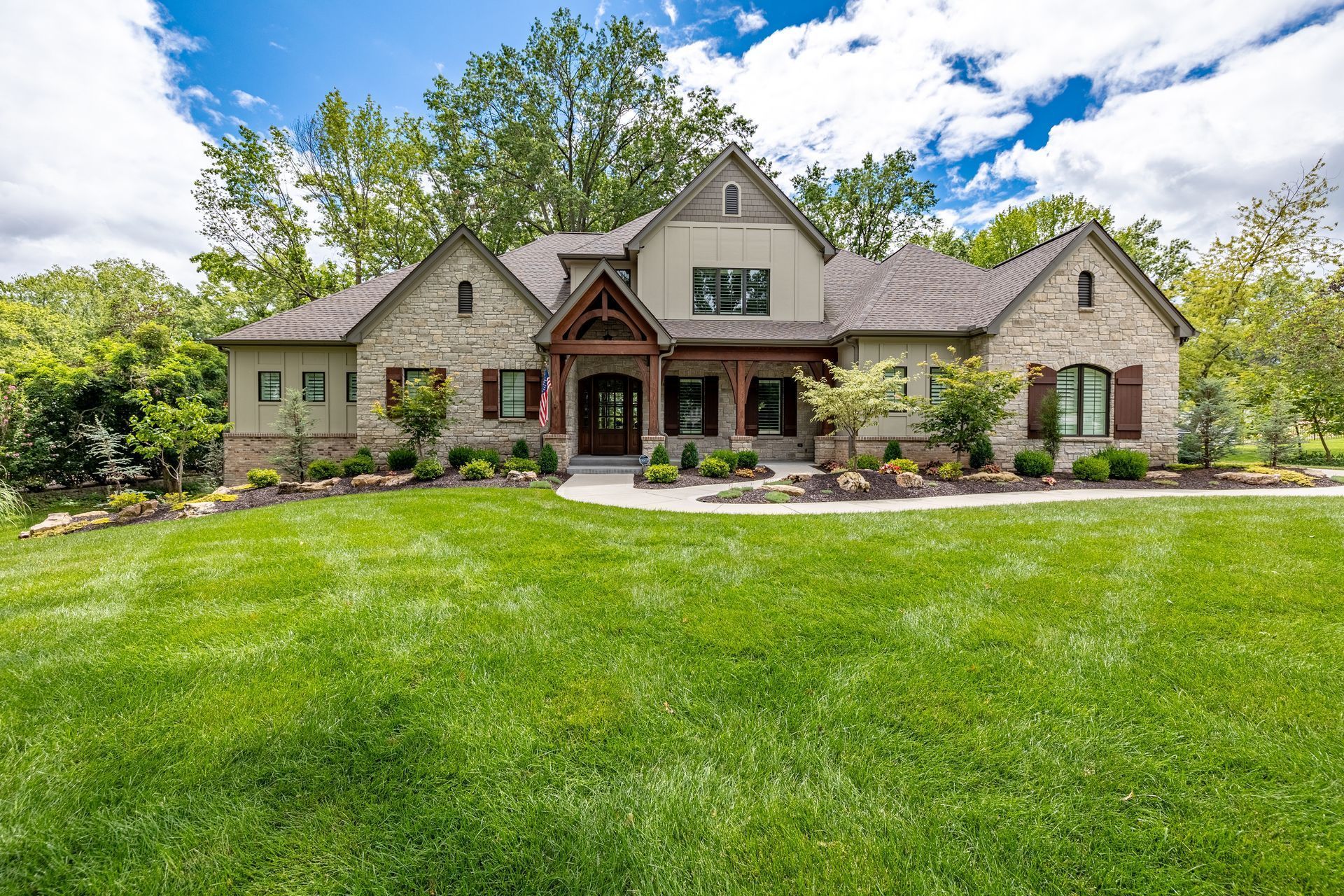Large stone house with a green lawn, trees, and a front porch under a partly cloudy sky