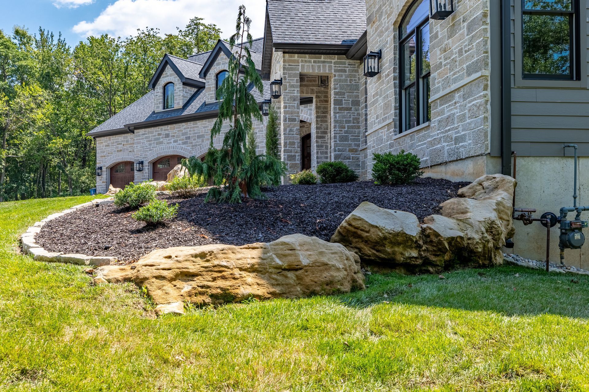 Stone house exterior with landscaped front yard, large rocks, and green lawn under sunny skies