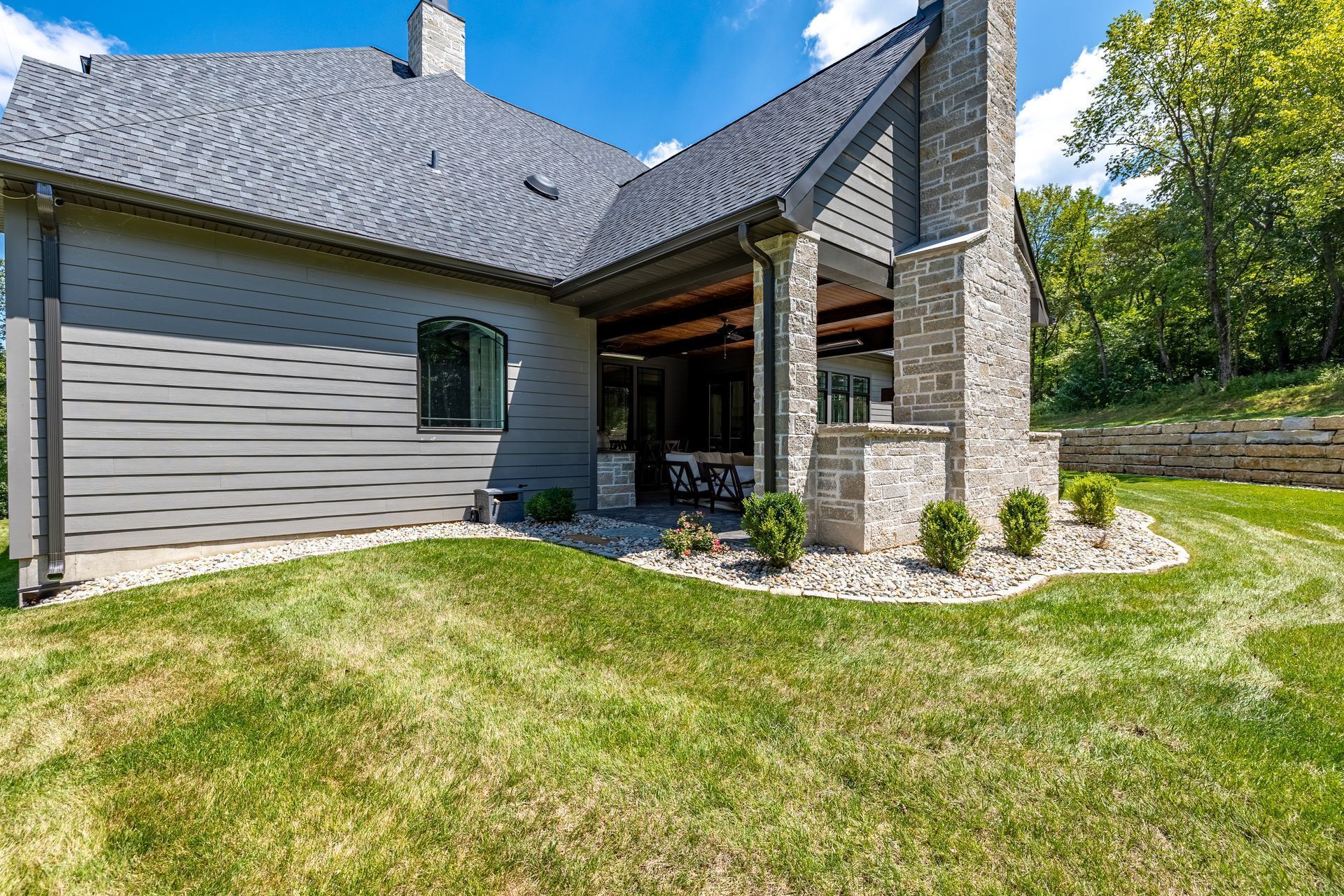 Modern house exterior with stone porch, gray siding, and a sloped green lawn under a blue sky