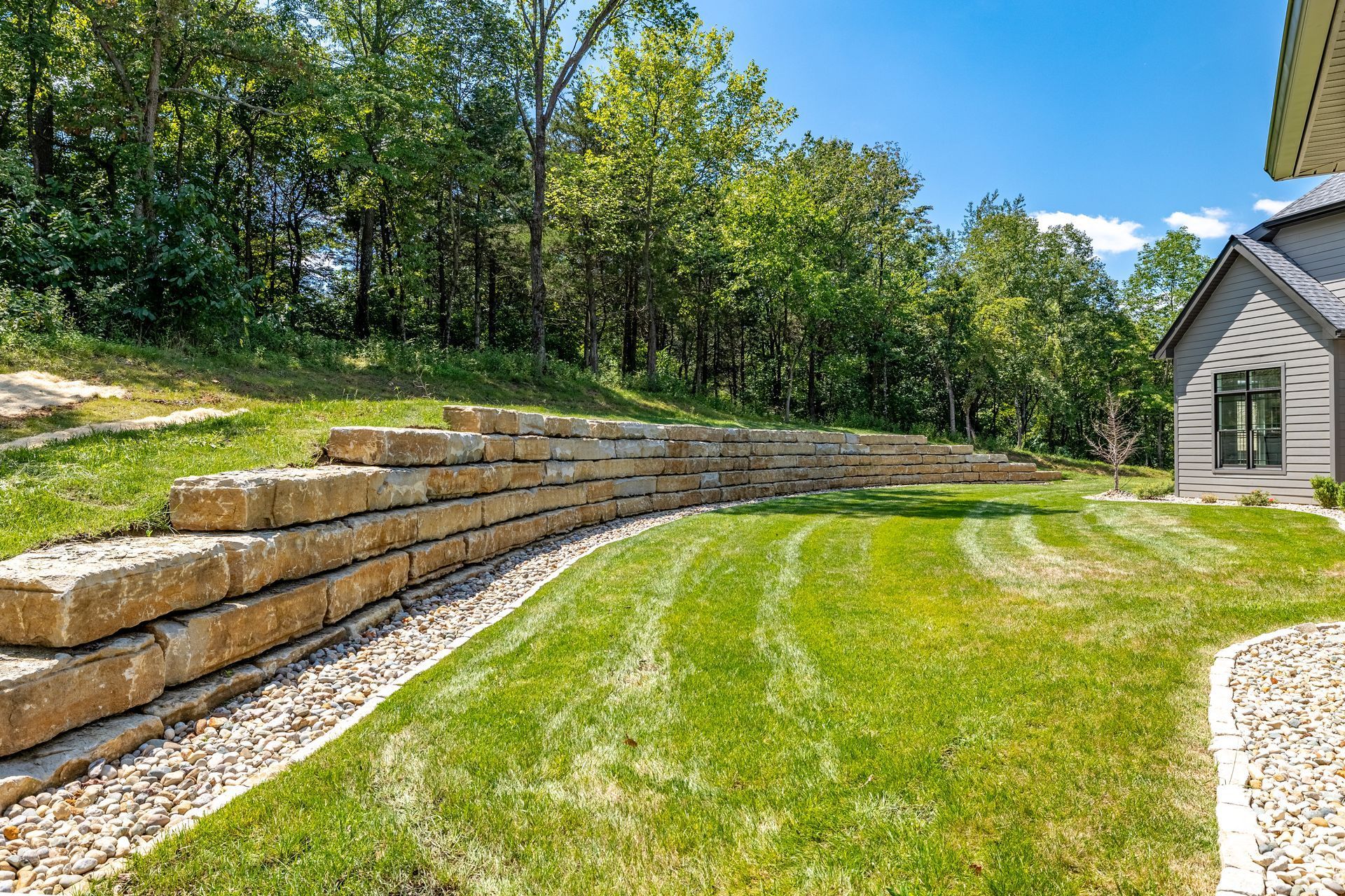Terraced stone retaining wall beside a green lawn and gravel path next to a house, with trees in the background