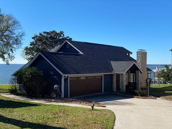 Blue house with garage, chimney, and dark roof sits by water on a sunny day.