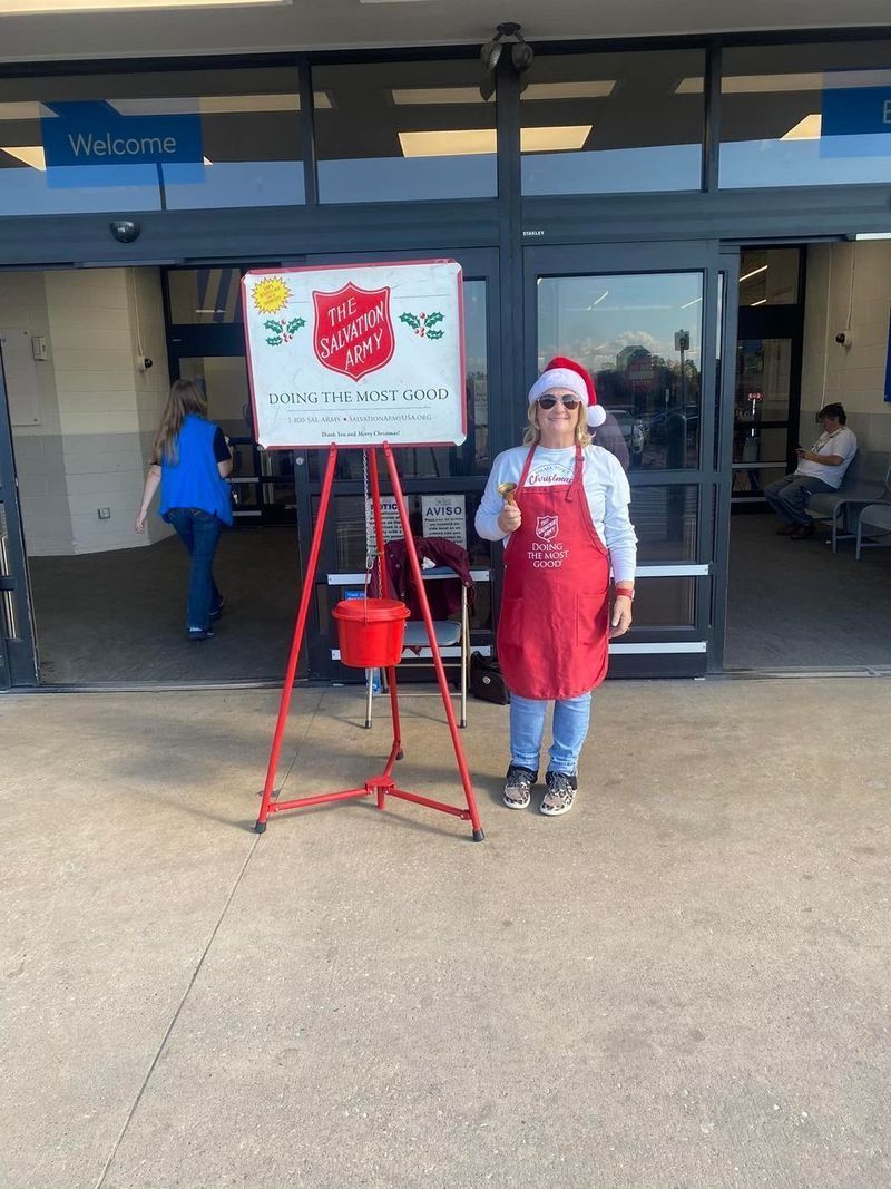 Person in Santa hat and apron rings bell for Salvation Army outside store entrance.