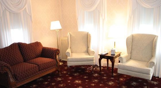 Living room with red patterned sofa, two cream wingback chairs, small table, floor lamp, and white curtains.