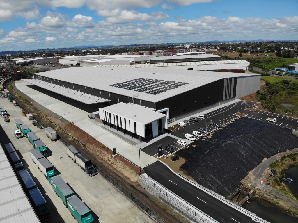 Aerial view of a large white industrial building beside a road with parked trucks and a dark roof area