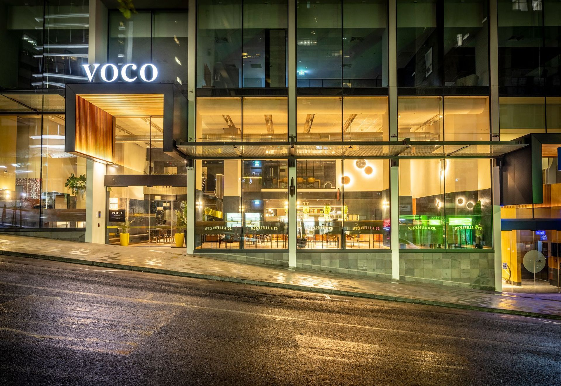 VOCO hotel entrance at night, with glass facade, warm interior lights, and wet street reflections