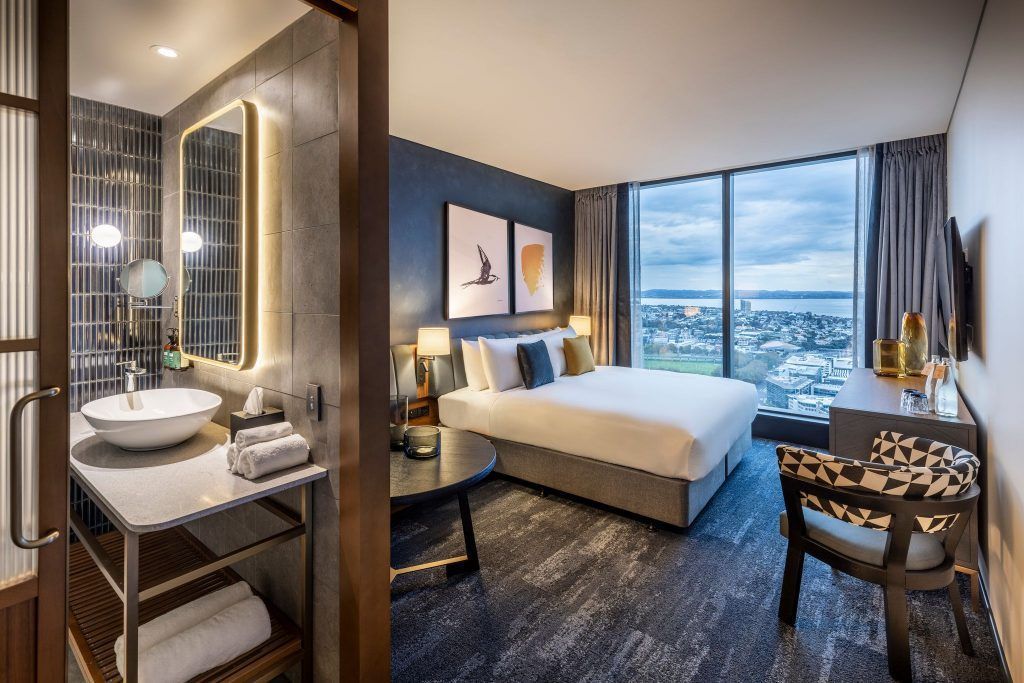Modern hotel room with king bed, desk, and city view through large windows