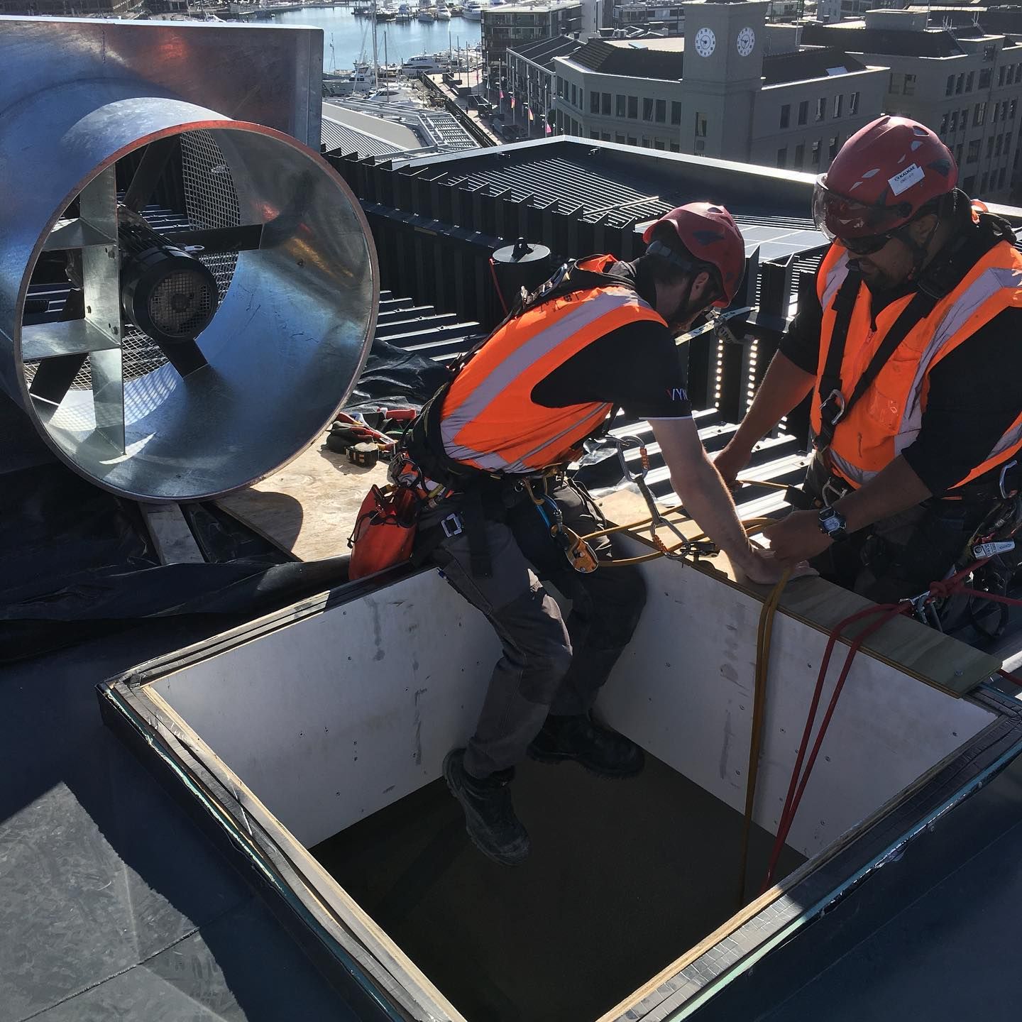 Two workers in orange safety vests on a rooftop near ventilation equipment and an open hatch.