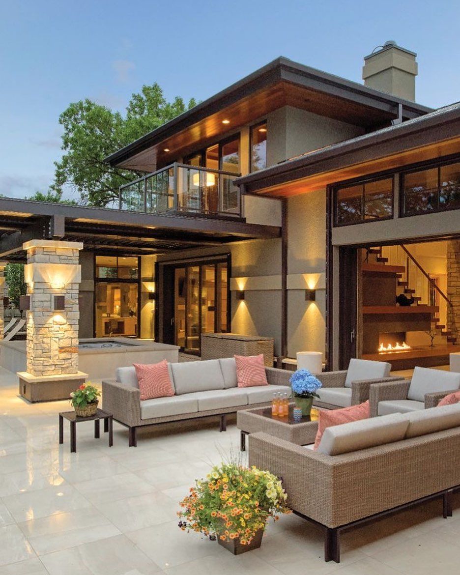 Modern luxury patio with wicker seating, cushions, and warm-lit stone house at dusk