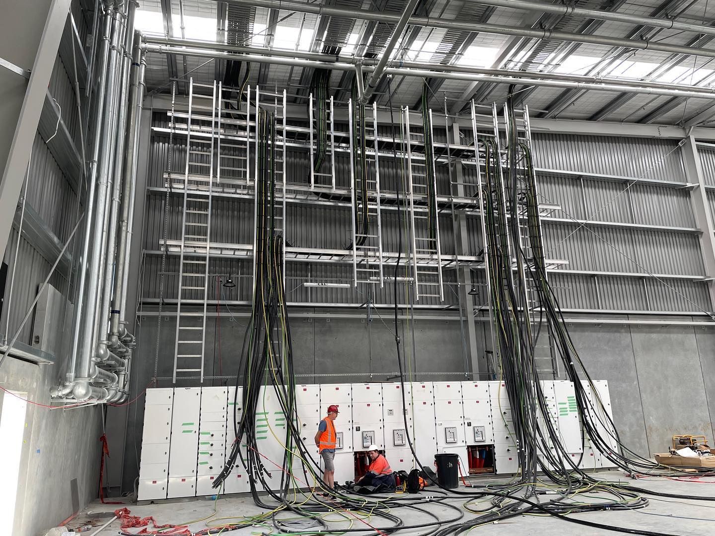 Workers in orange gear beneath tall hanging cables inside an industrial warehouse.