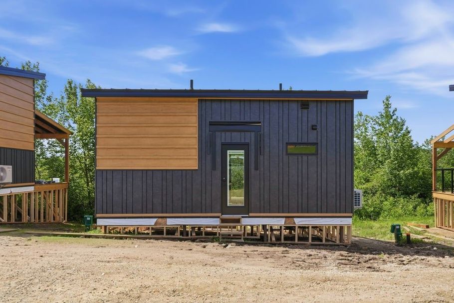 Modern small cabin with dark gray and wood siding, centered door, in a sunny rural setting.