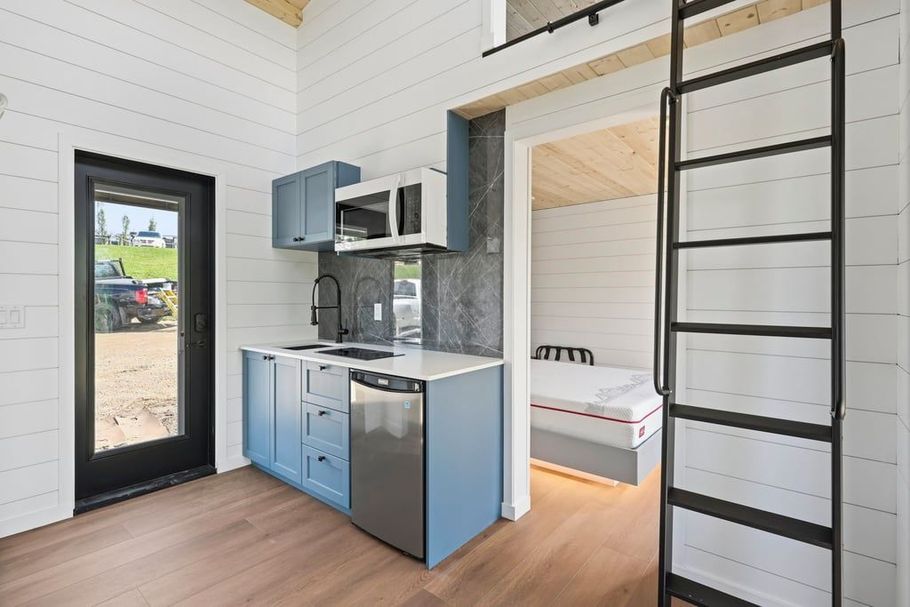 Compact tiny-house kitchen with blue cabinets, loft bed, and ladder in a bright white interior