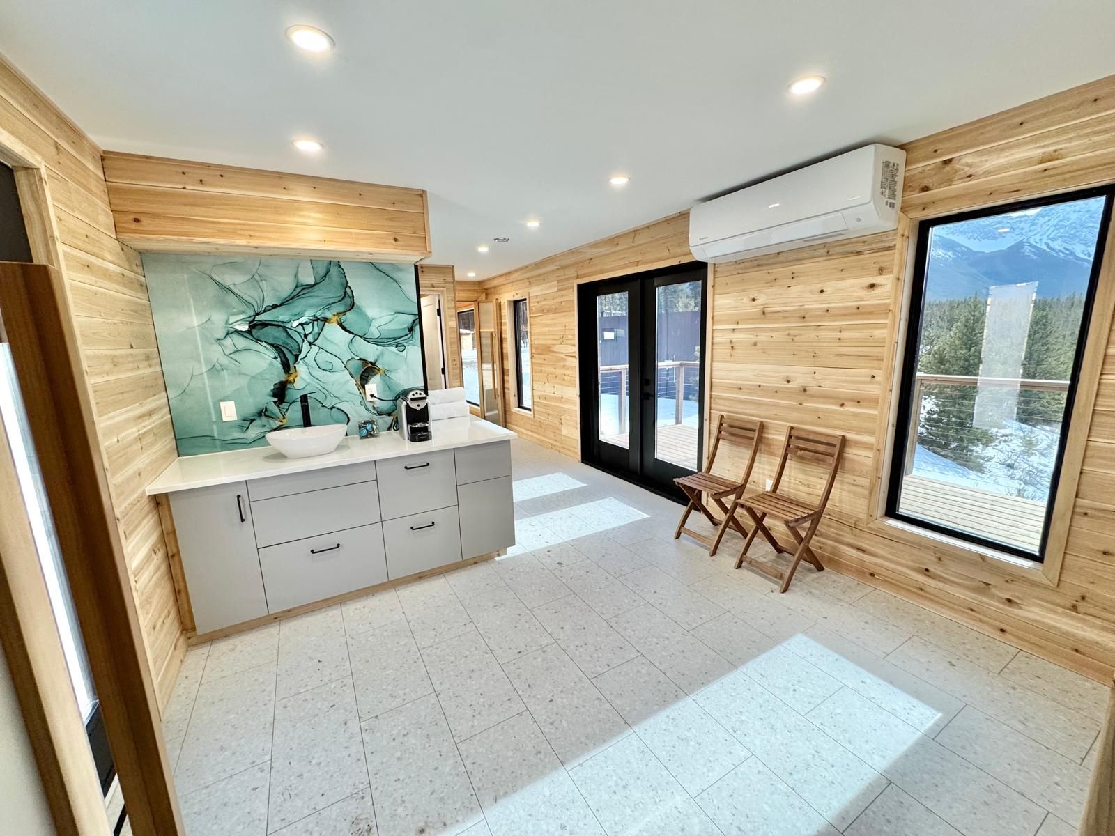 Modern bright room with wood walls, a kitchenette, abstract wall art, two chairs, and large windows.