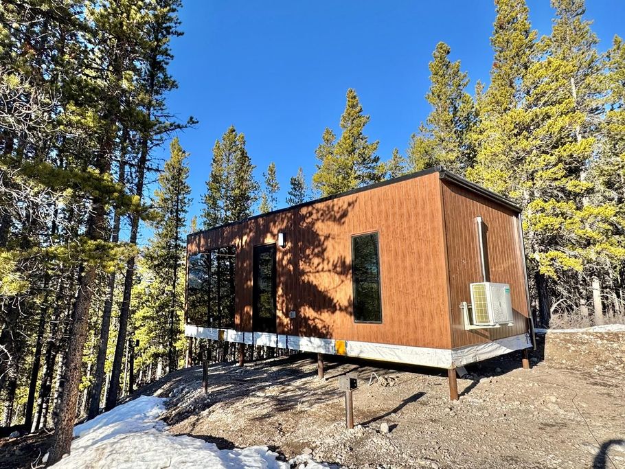 Small brown cabin on stilts in a snowy pine forest under a bright blue sky.