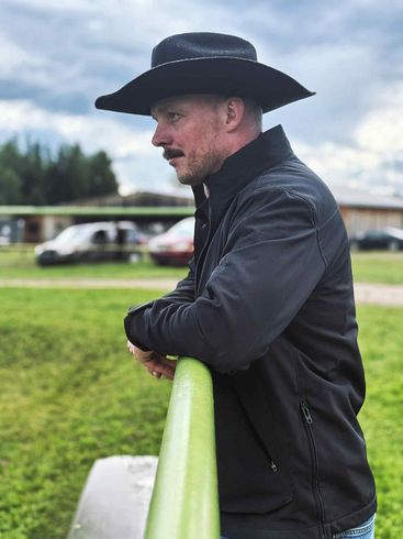 Man in a black hat and jacket leaning on a green fence at a racetrack
