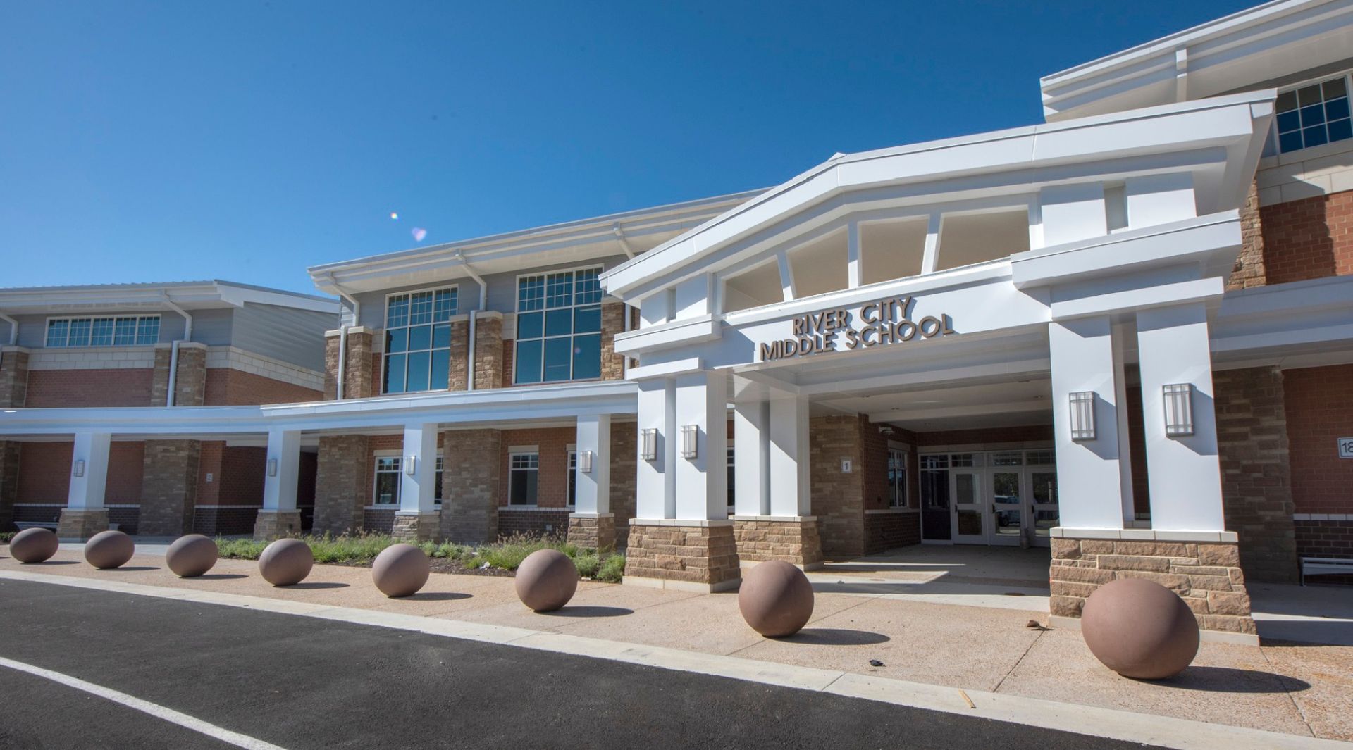 Exterior of a modern building with white columns, glass windows, and stone bollards under a clear blue sky