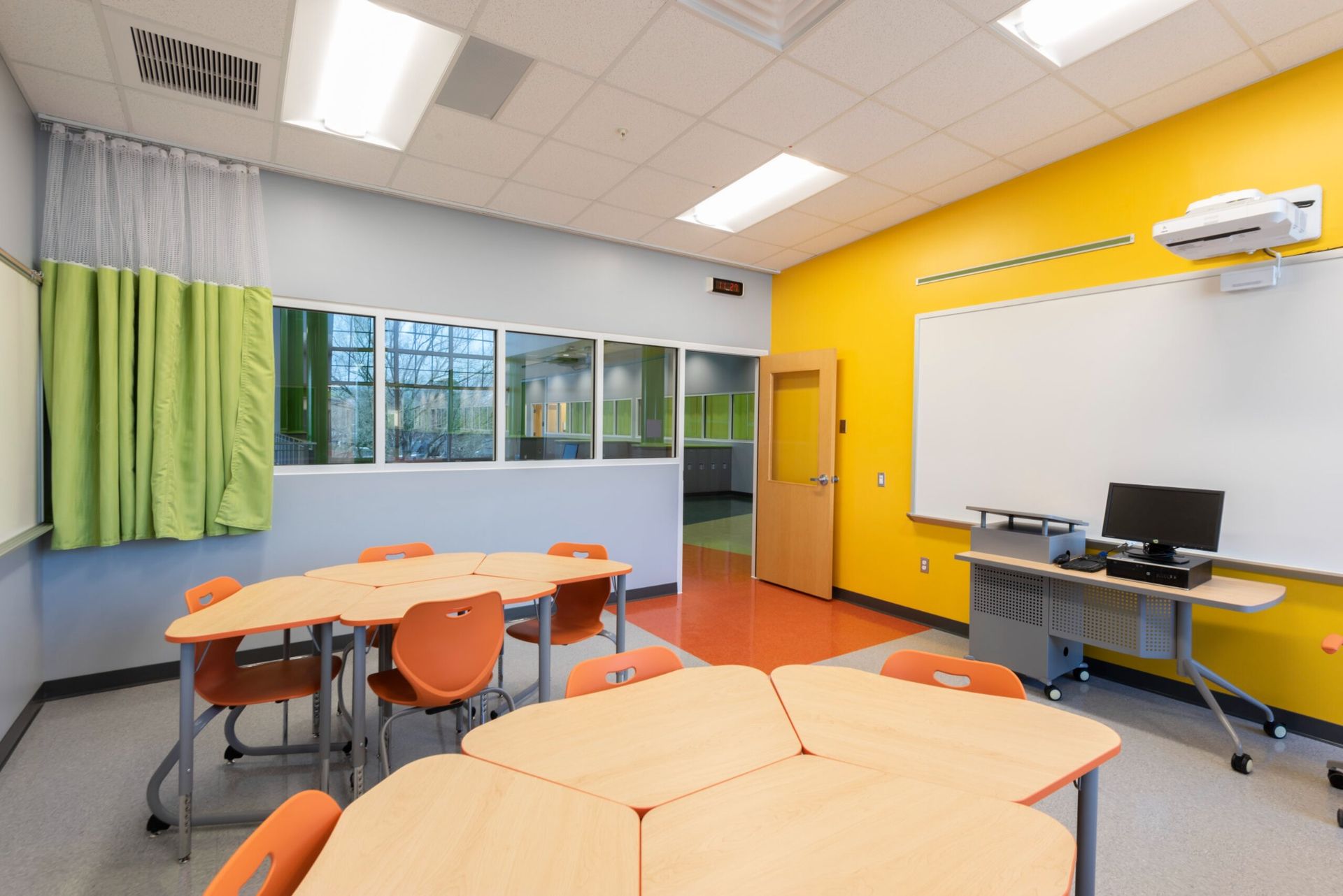 Bright classroom with orange chairs, wooden tables, large windows, and a desk against a yellow wall.