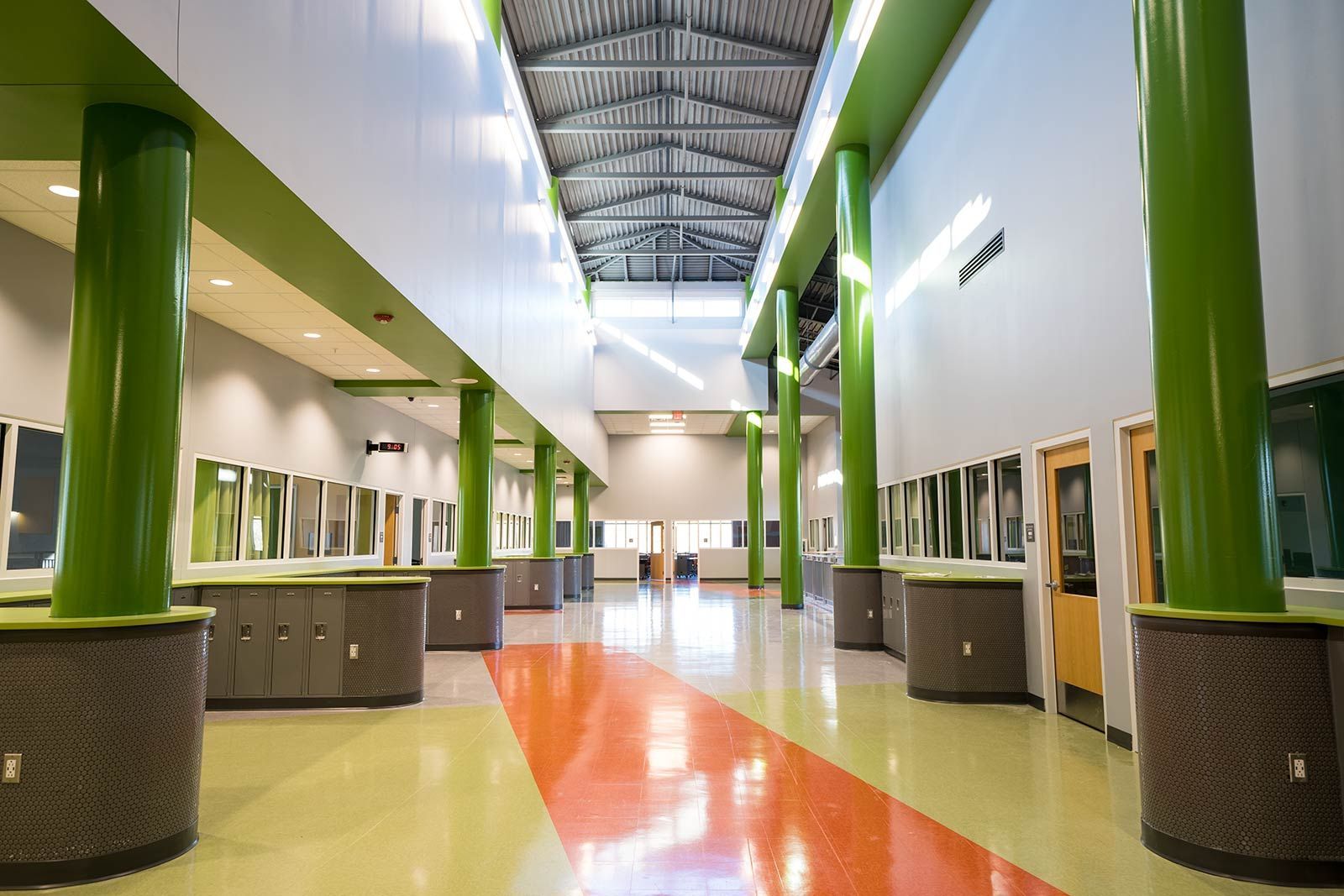 Bright school hallway with green columns, orange floor stripe, and windows along both sides.